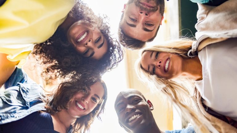 A diverse group of six young adults smiles as they huddle together, looking down at the camera from a low-angle perspective, creating a sense of unity and shared joy.